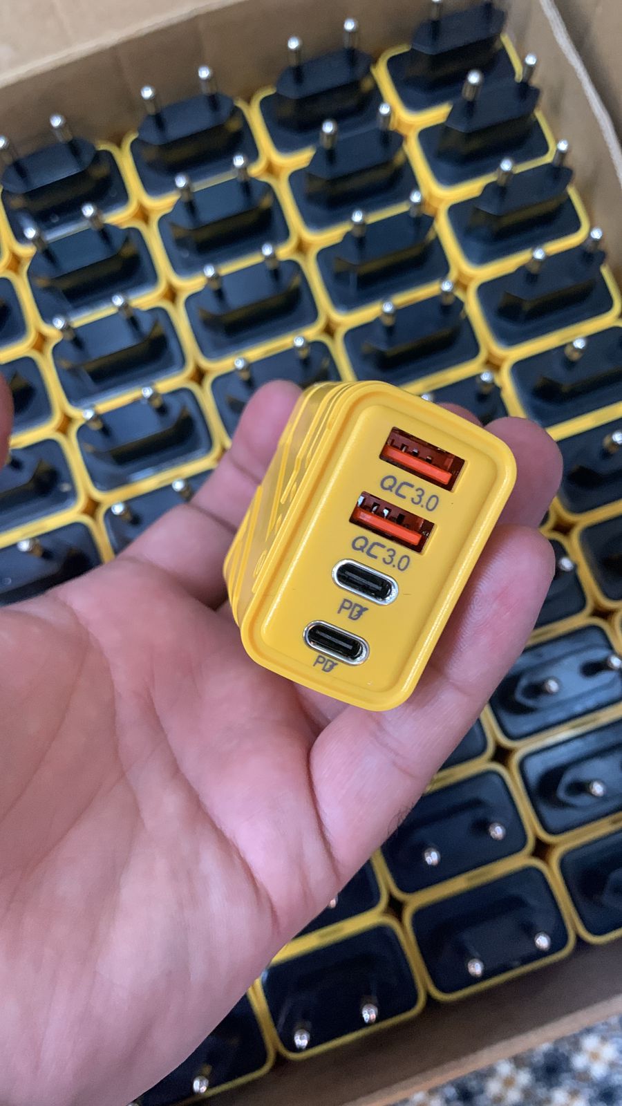 Yellow USB charger with multiple ports held in a hand, with a grid of similar chargers in the background.