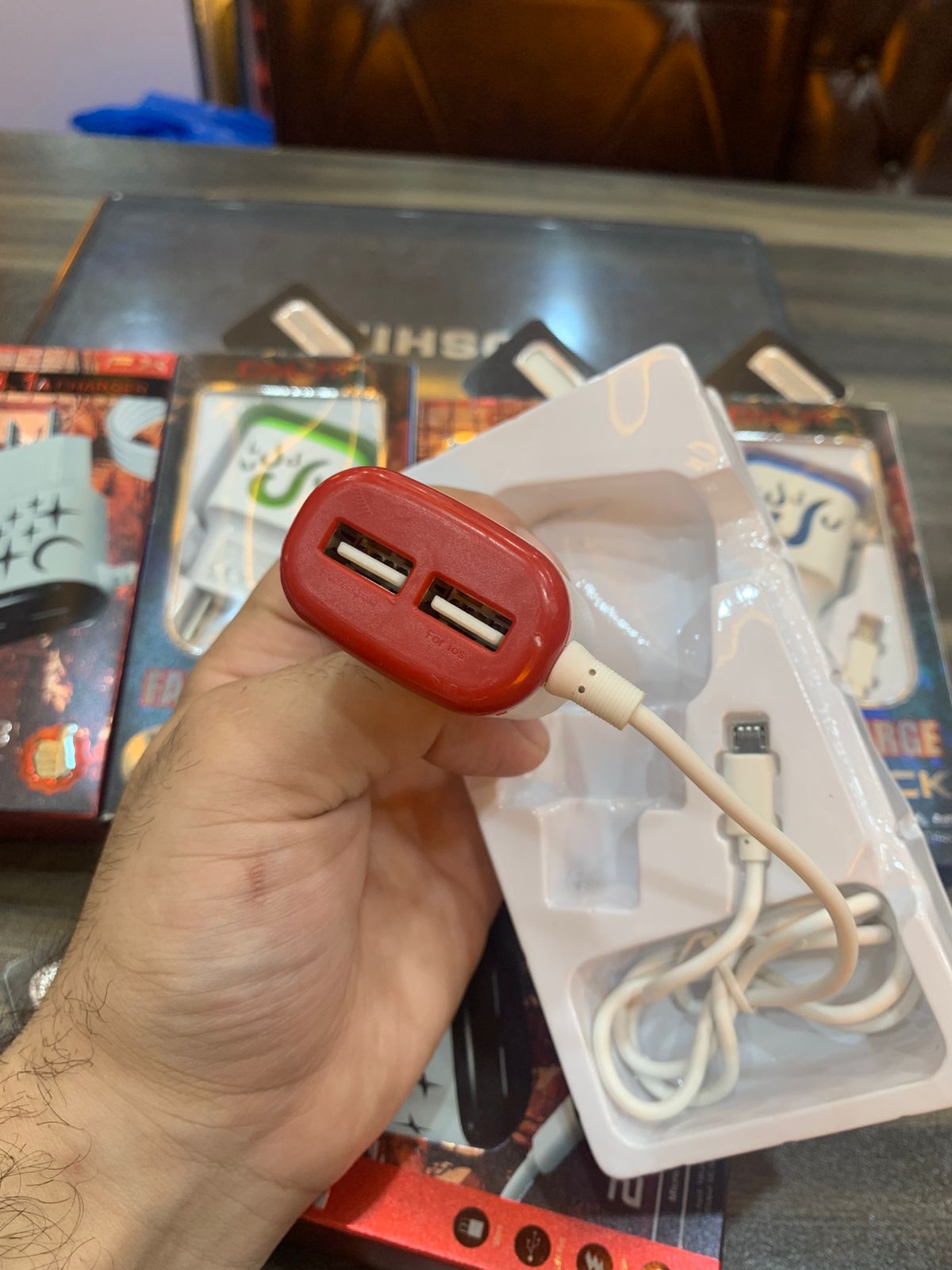 Red USB charger with a cable held by a hand, with a blurred background of gaming merchandise.